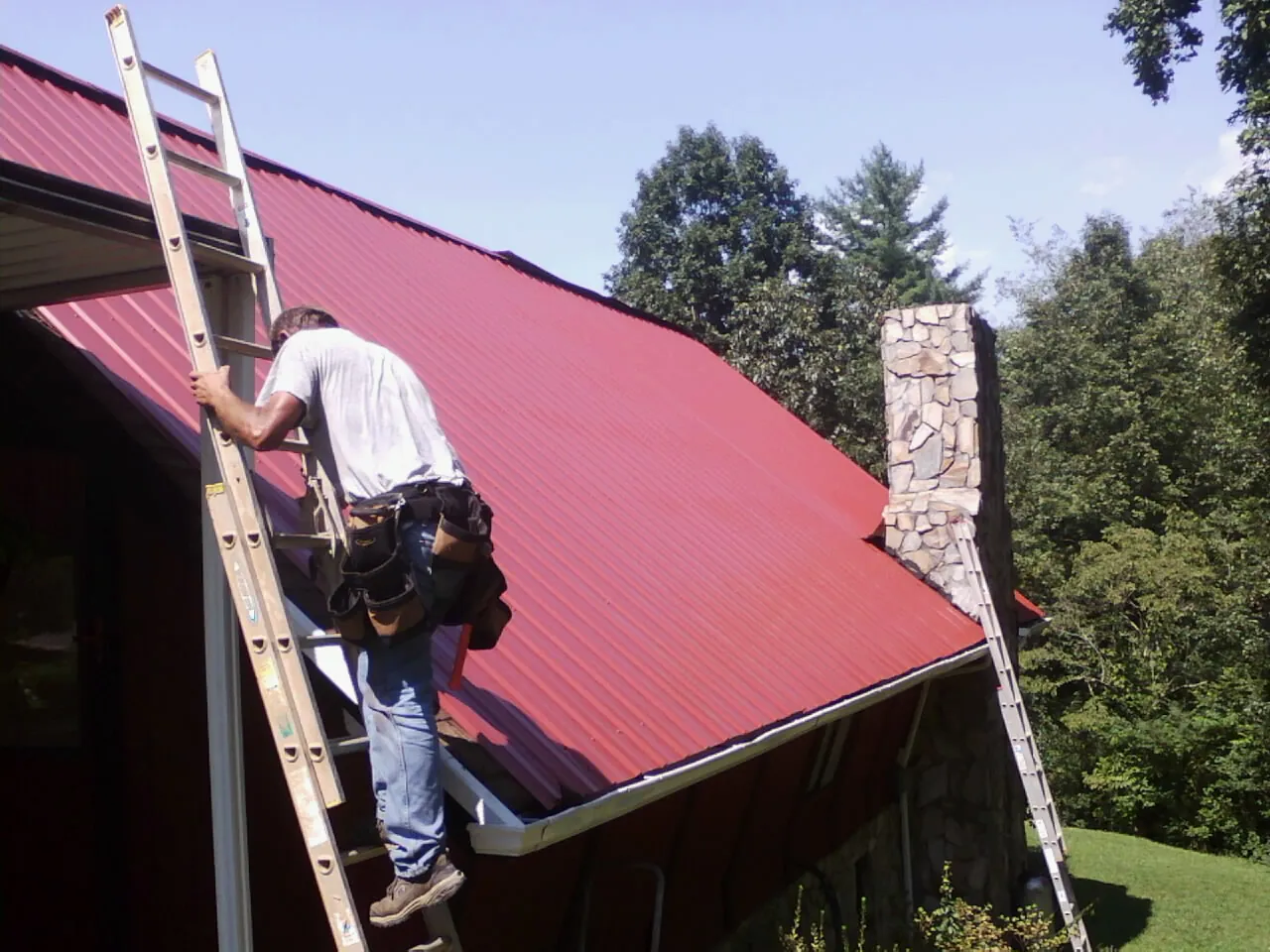 Roofer on a ladder beside a red metal roof and stone chimney — Soffit & Fascia Repair by Roofing Brothers in Waddy